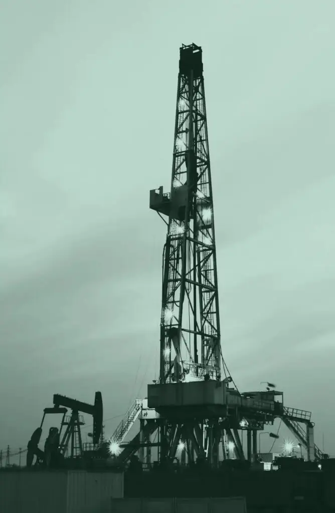 Illuminated oil drilling rig at dusk with pumpjack and workers in the foreground, representing the energy sector.